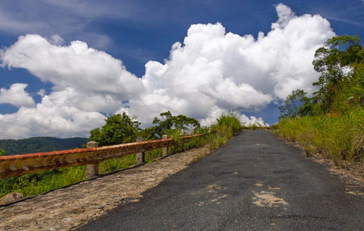 A road rises from the bottom of the frame towards the horizon under large white clouds